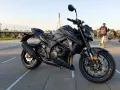 Czarny motocykl Suzuki GSX-S 750 z niebieskimi akcentami na felgach i owiewkach, zaparkowany na placu z widocznym w tle mostem i ludźmi.
