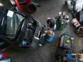 Warsztat samochodowy z dwoma mechanikami przy otwartej masce czarnego auta i narzędziami.
