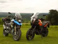 Dwa motocykle turystyczne, w tym pomarańczowy KTM 1290 Super Adventure, stoją na trawie z widokiem na zielone wzgórza.