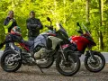 Dwóch motocyklistów z kaskiem na siedzeniu obok dwóch motocykli Ducati Multistrada, szary i czerwony, na tle leśnej drogi.