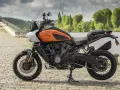 Pomarańczowy Harley-Davidson Pan America gotowy na przygodę, z mostem i zielenią w tle.