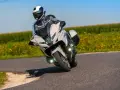 Motocyklista w kasku i kombinezonie pochyla się na zakręcie na białym motocyklu BMW R 1250 RT, przemierzając malowniczą drogę wśród pól.