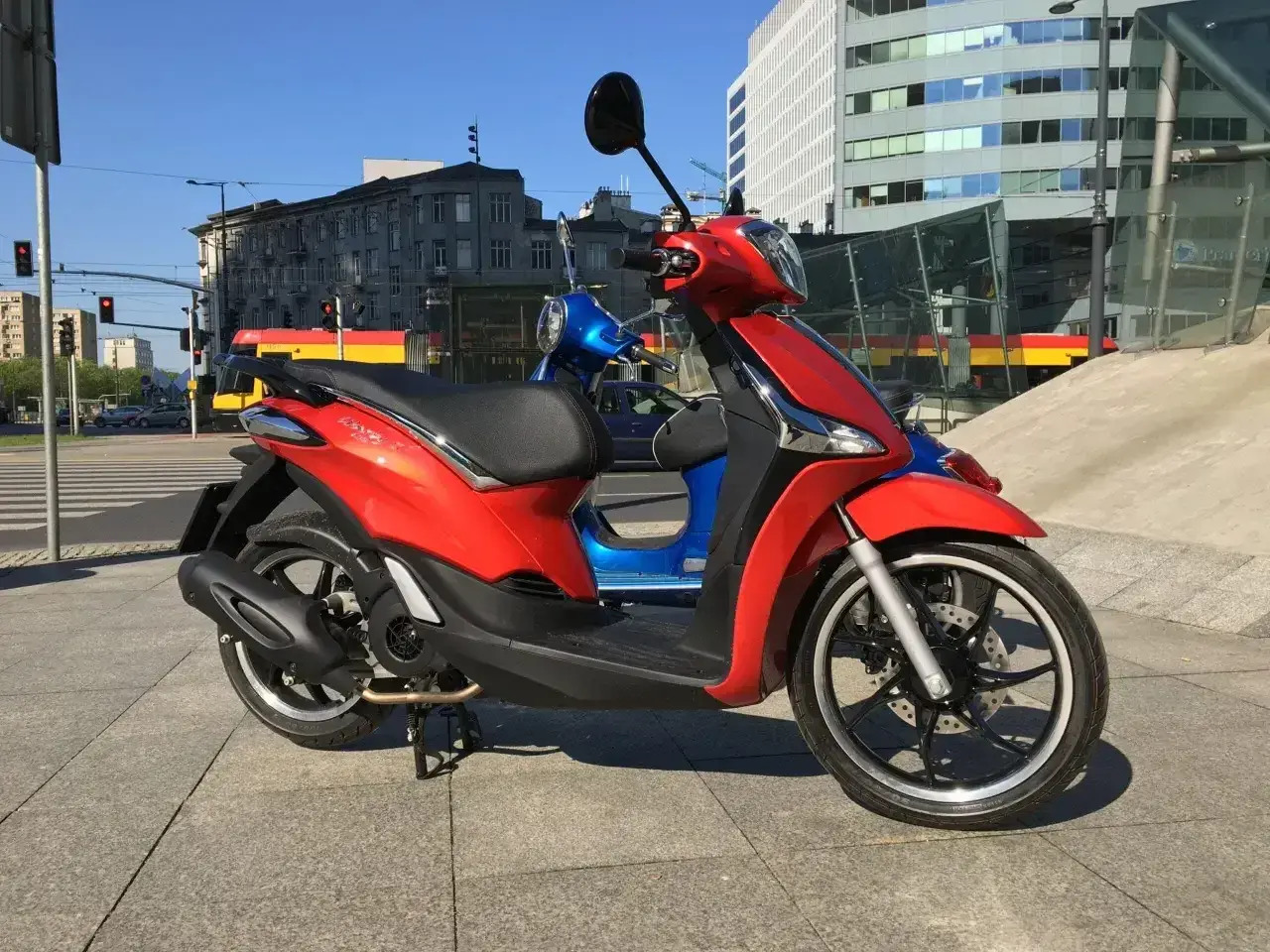 Czerwony Piaggio Liberty 125, obok niebieskiego, na tle miasta. Idealny do sprawdzenia piaggio liberty 125 dane techniczne.