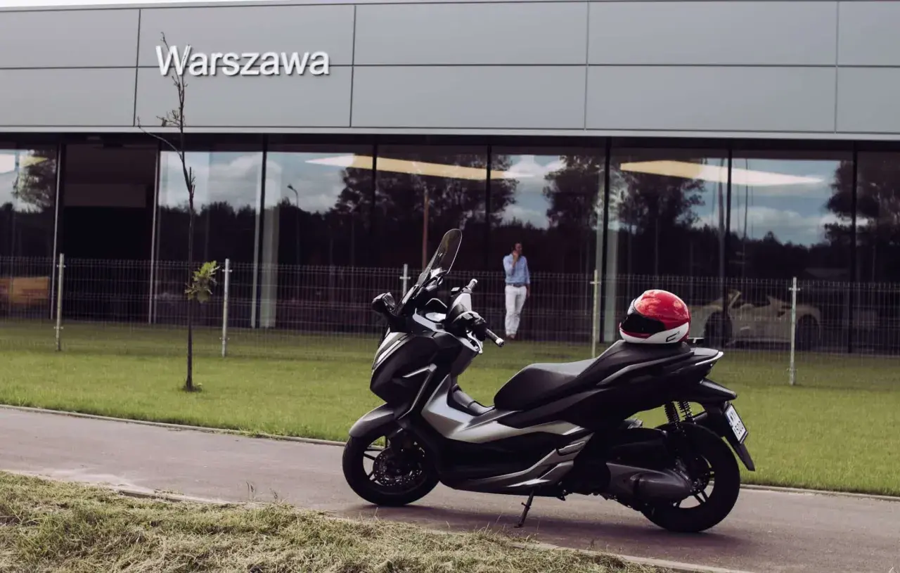 Elegancki skuter Honda Forza 300 zaparkowany przed budynkiem z napisem "Warszawa". Czerwony kask leży na siedzeniu.