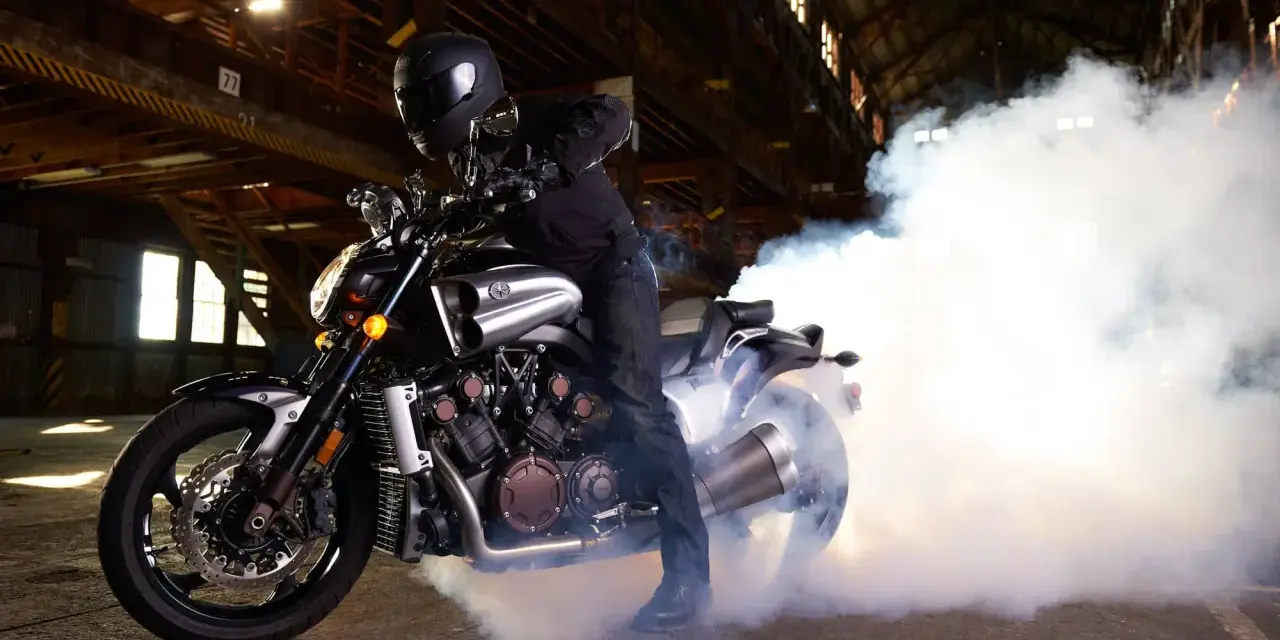 Motocyklista w kasku na Yamasze V-Max wykonuje burnout, tworząc kłęby dymu w opuszczonej hali.