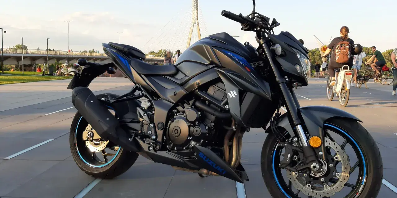 Czarny motocykl Suzuki GSX-S 750 z niebieskimi akcentami na felgach i owiewkach, zaparkowany na placu z widocznym w tle mostem i ludźmi.