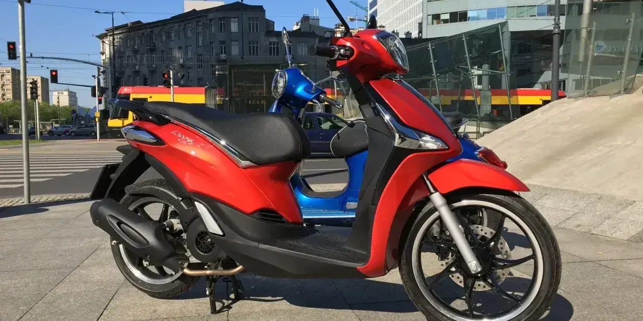 Czerwony Piaggio Liberty 125, obok niebieskiego, na tle miasta. Idealny do sprawdzenia piaggio liberty 125 dane techniczne.