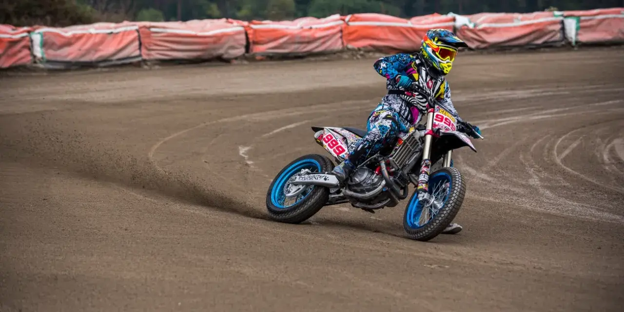 Motocyklista w kolorowym stroju śmiga po piaszczystym torze **flat track**, pochylony w zakręcie.