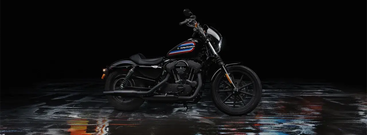 Czarny Harley-Davidson Iron 1200 z kolorowym paskiem na baku, stojący na mokrej, błyszczącej nawierzchni.