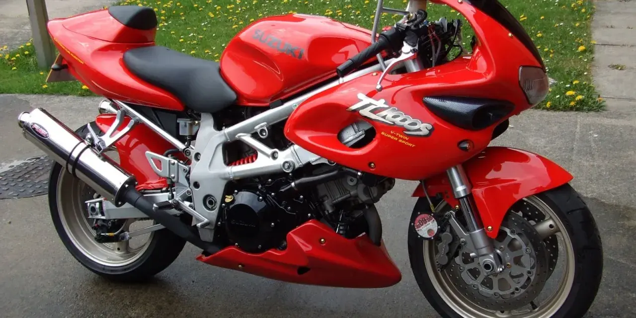 Czerwony motocykl Suzuki TL1000 z napisem "V-TWIN SUPER SPORT" na owiewce, zaparkowany na mokrej nawierzchni obok trawnika.
