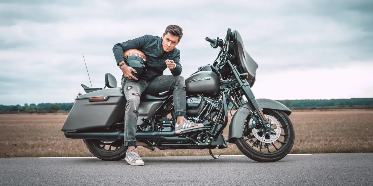 Mężczyzna w czarnej kurtce i podartych jeansach siedzi na motocyklu Harley-Davidson Street Glide, trzymając kask.