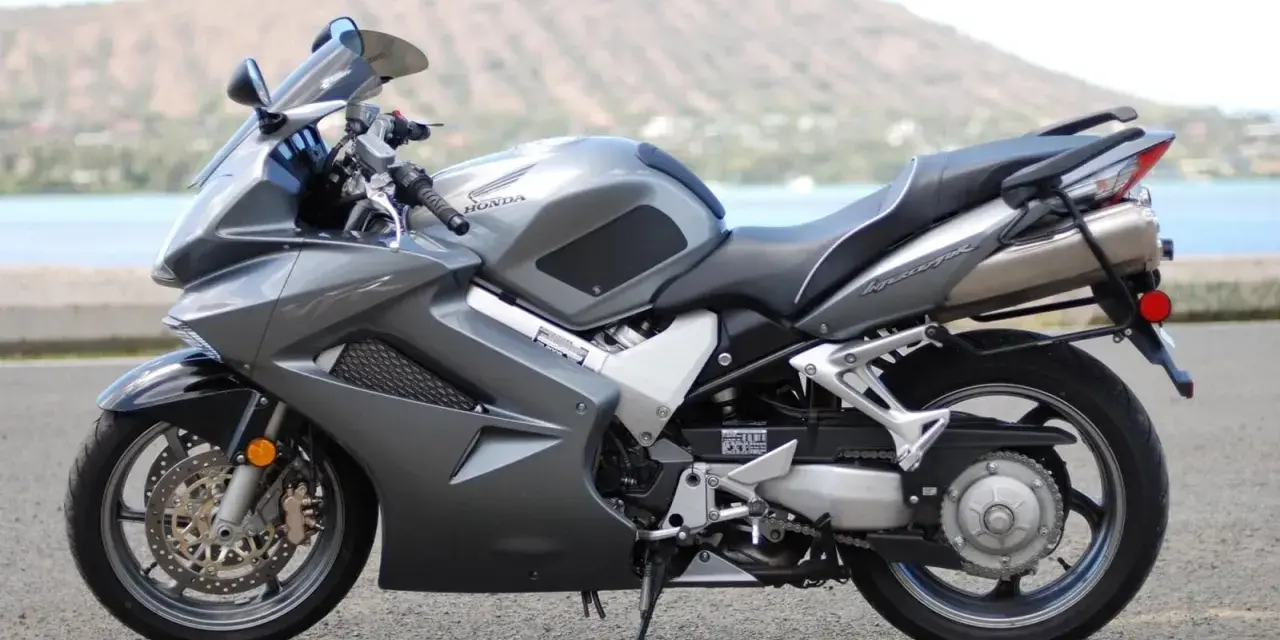 Szary motocykl Honda VFR 800 z widokiem na wodę i wzgórza w tle.