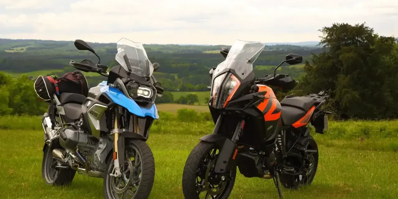 Dwa motocykle turystyczne, w tym pomarańczowy KTM 1290 Super Adventure, stoją na trawie z widokiem na zielone wzgórza.