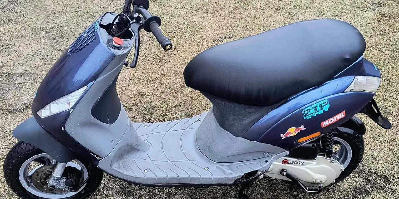Niebieski skuter z naklejkami Red Bull i Motul na tle trawy.