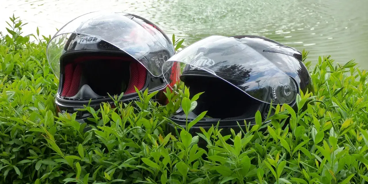Dwa kaski motocyklowe w zielonych liściach, z wodą w tle.
