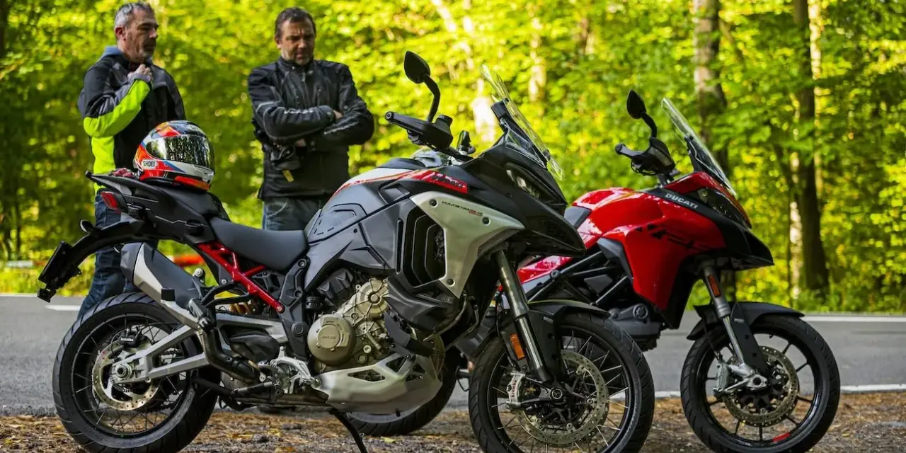 Dwóch motocyklistów z kaskiem na siedzeniu obok dwóch motocykli Ducati Multistrada, szary i czerwony, na tle leśnej drogi.