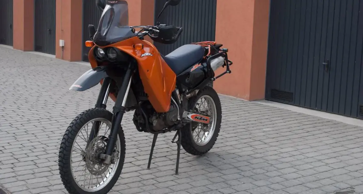 Pomarańczowy motocykl KTM 640 Adventure stoi na brukowanym podjeździe przed garażami.