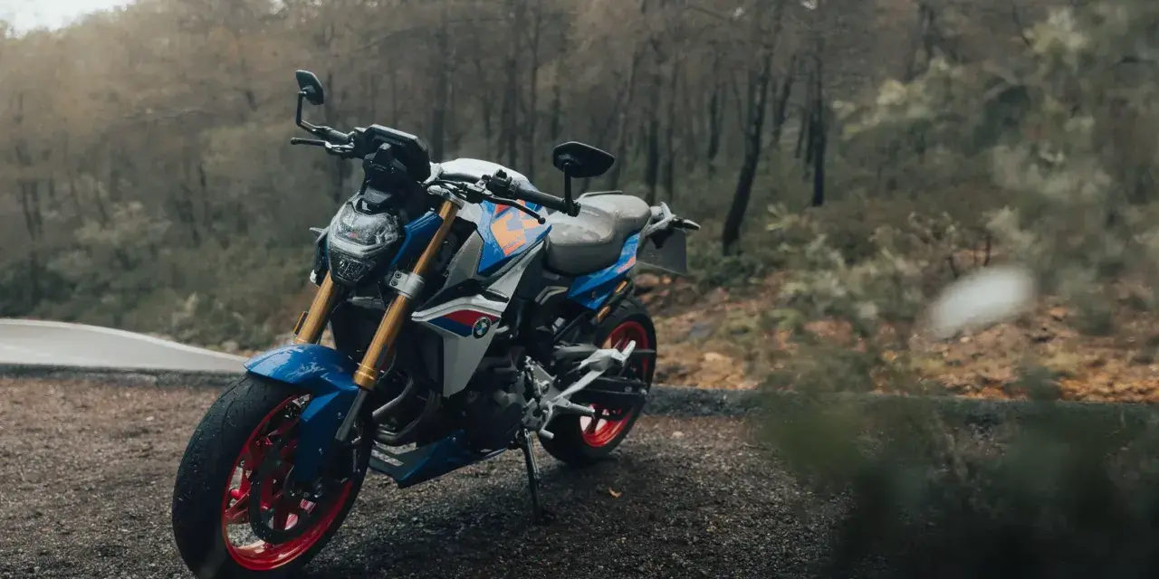 Niebiesko-biały motocykl BMW F900R z czerwonymi felgami na tle jesiennego lasu.
