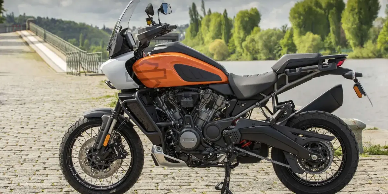 Pomarańczowy Harley-Davidson Pan America gotowy na przygodę, z mostem i zielenią w tle.