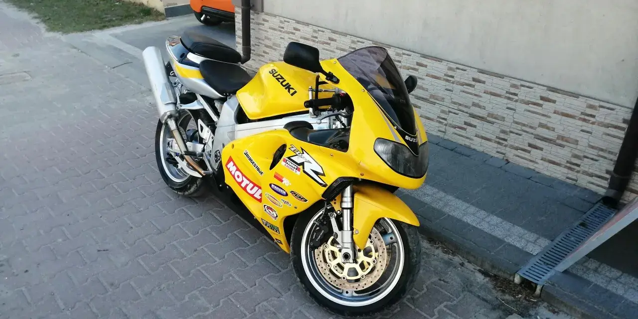 Żółty motocykl Suzuki TL1000R z naklejkami sponsorów, zaparkowany na chodniku.