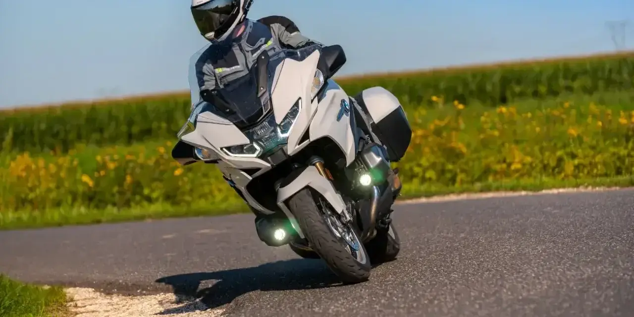 Motocyklista w kasku i kombinezonie pochyla się na zakręcie na białym motocyklu BMW R 1250 RT, przemierzając malowniczą drogę wśród pól.