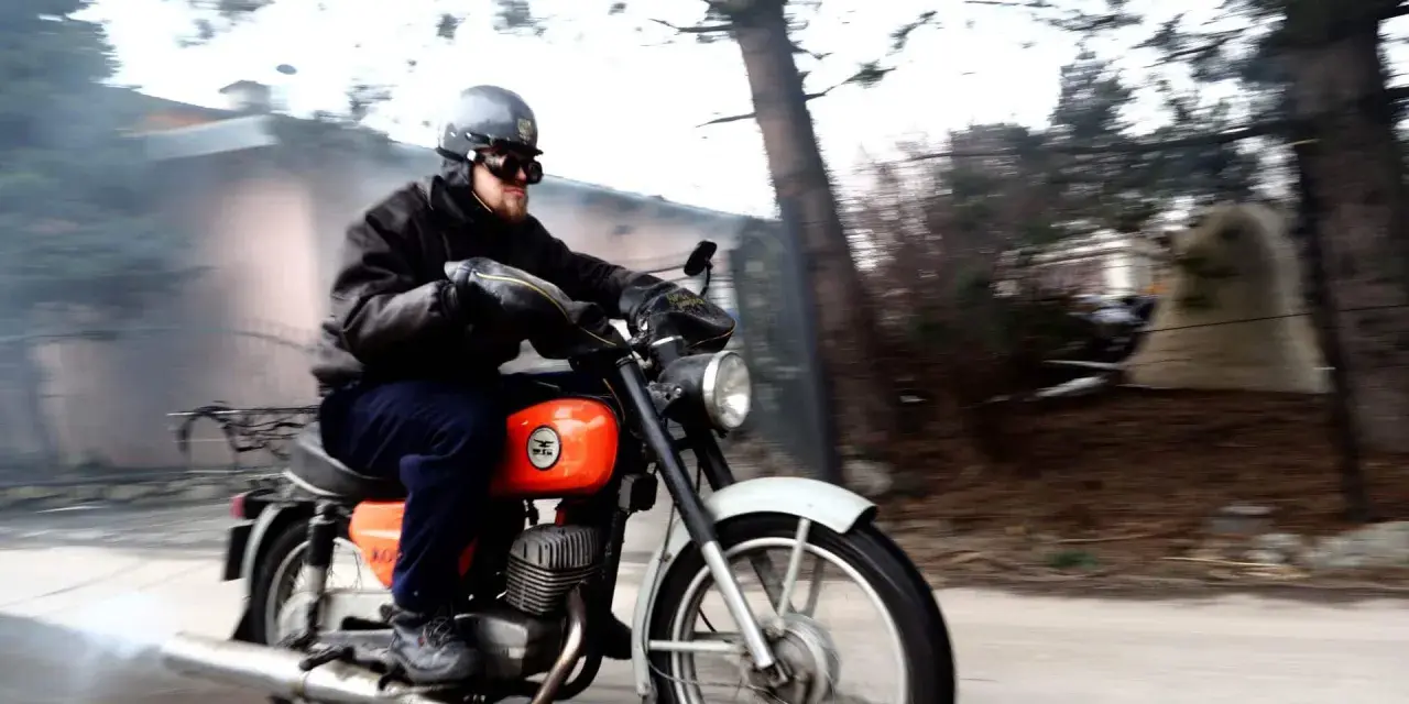 Mężczyzna w kasku i goglach pędzi na pomarańczowym motocyklu WSK 125. Widać dym z rury wydechowej, sugerujący dużą prędkość maksymalną.