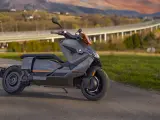 Szary skuter elektryczny BMW CE 04 na tle malowniczego krajobrazu z mostem i wzgórzami.