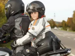 Uśmiechnięte dziecko na motocyklu, siedzące przed dorosłym, w kasku i kurtce motocyklowej.