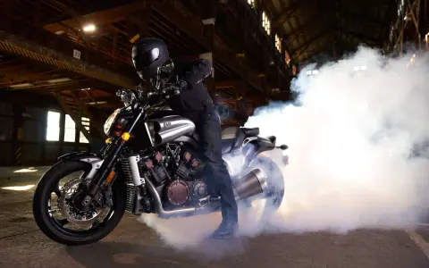 Motocyklista w kasku na Yamasze V-Max wykonuje burnout, tworząc kłęby dymu w opuszczonej hali.