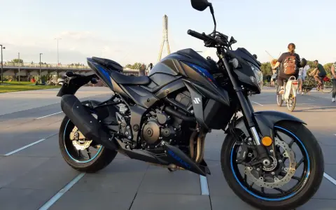 Czarny motocykl Suzuki GSX-S 750 z niebieskimi akcentami na felgach i owiewkach, zaparkowany na placu z widocznym w tle mostem i ludźmi.