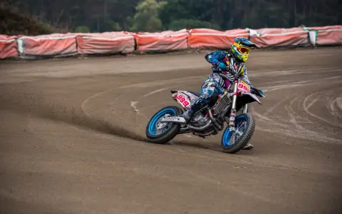 Motocyklista w kolorowym stroju śmiga po piaszczystym torze **flat track**, pochylony w zakręcie.