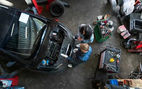 Warsztat samochodowy z dwoma mechanikami przy otwartej masce czarnego auta i narzędziami.
