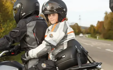 Uśmiechnięte dziecko na motocyklu, siedzące przed dorosłym, w kasku i kurtce motocyklowej.