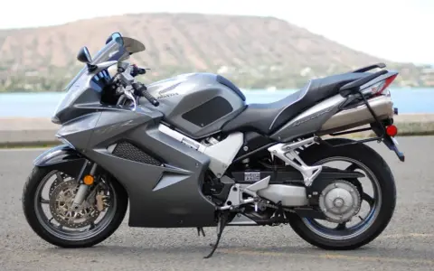 Szary motocykl Honda VFR 800 z widokiem na wodę i wzgórza w tle.