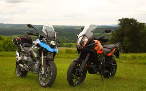 Dwa motocykle turystyczne, w tym pomarańczowy KTM 1290 Super Adventure, stoją na trawie z widokiem na zielone wzgórza.