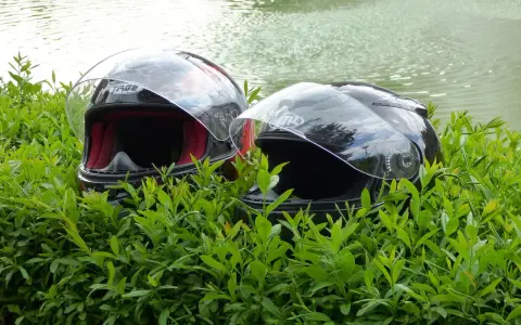 Dwa kaski motocyklowe w zielonych liściach, z wodą w tle.