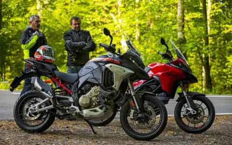 Dwóch motocyklistów z kaskiem na siedzeniu obok dwóch motocykli Ducati Multistrada, szary i czerwony, na tle leśnej drogi.