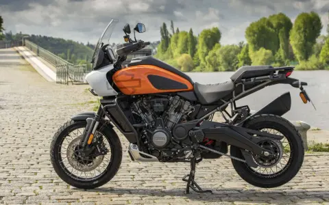 Pomarańczowy Harley-Davidson Pan America gotowy na przygodę, z mostem i zielenią w tle.