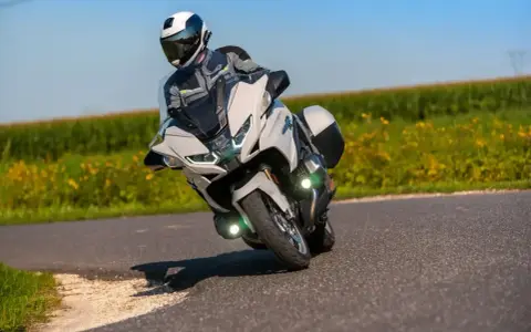 Motocyklista w kasku i kombinezonie pochyla się na zakręcie na białym motocyklu BMW R 1250 RT, przemierzając malowniczą drogę wśród pól.