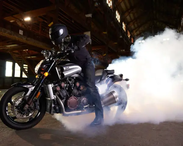 Motocyklista w kasku na Yamasze V-Max wykonuje burnout, tworząc kłęby dymu w opuszczonej hali.