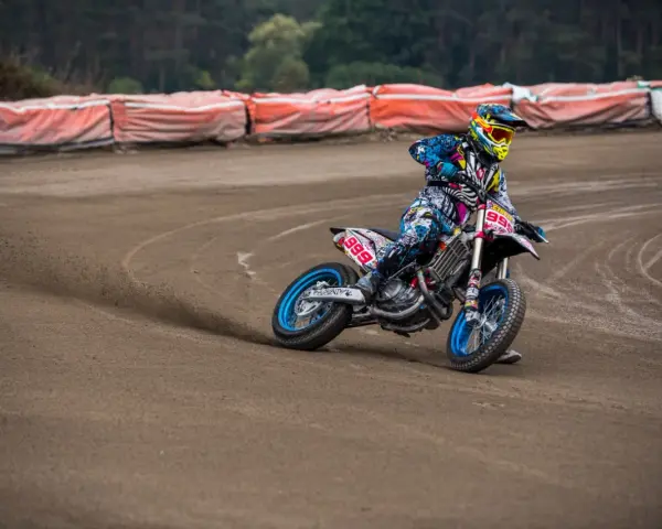 Motocyklista w kolorowym stroju śmiga po piaszczystym torze **flat track**, pochylony w zakręcie.