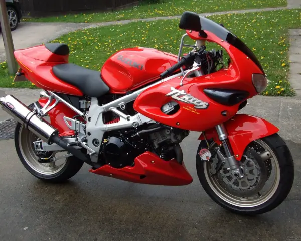 Czerwony motocykl Suzuki TL1000 z napisem "V-TWIN SUPER SPORT" na owiewce, zaparkowany na mokrej nawierzchni obok trawnika.