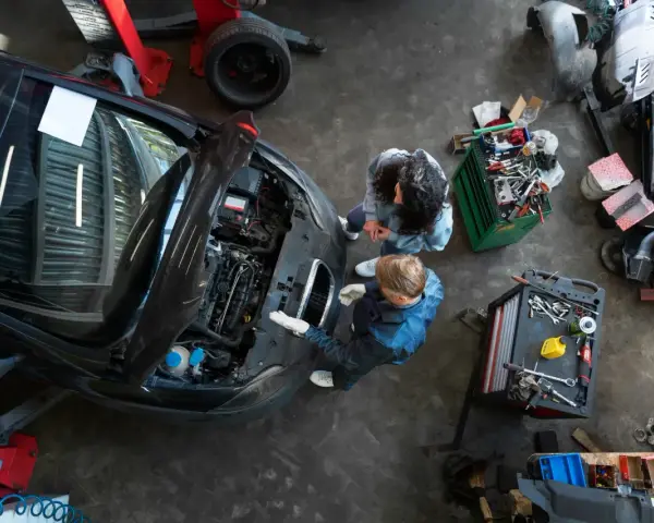 Warsztat samochodowy z dwoma mechanikami przy otwartej masce czarnego auta i narzędziami.