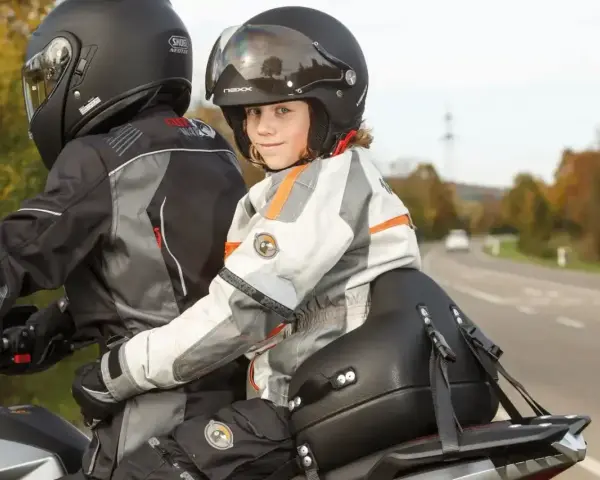Uśmiechnięte dziecko na motocyklu, siedzące przed dorosłym, w kasku i kurtce motocyklowej.