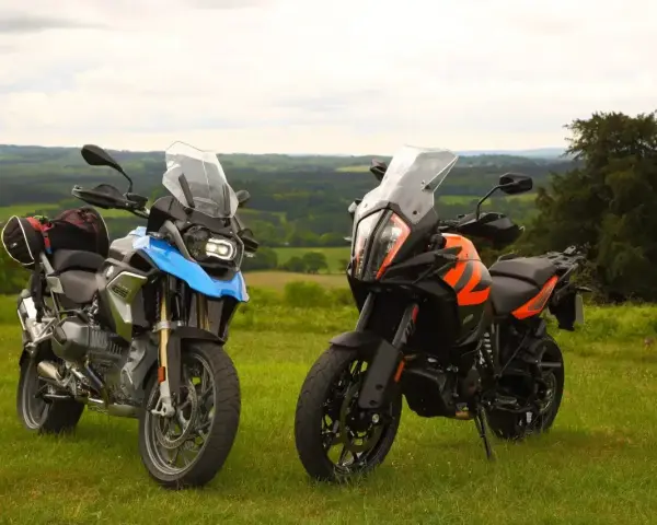 Dwa motocykle turystyczne, w tym pomarańczowy KTM 1290 Super Adventure, stoją na trawie z widokiem na zielone wzgórza.