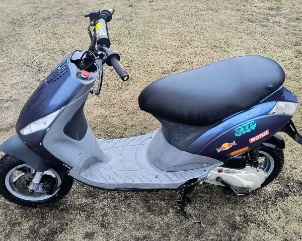 Niebieski skuter z naklejkami Red Bull i Motul na tle trawy.