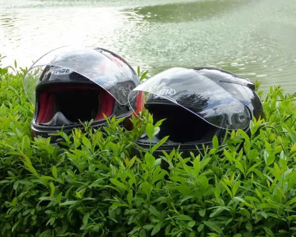 Dwa kaski motocyklowe w zielonych liściach, z wodą w tle.