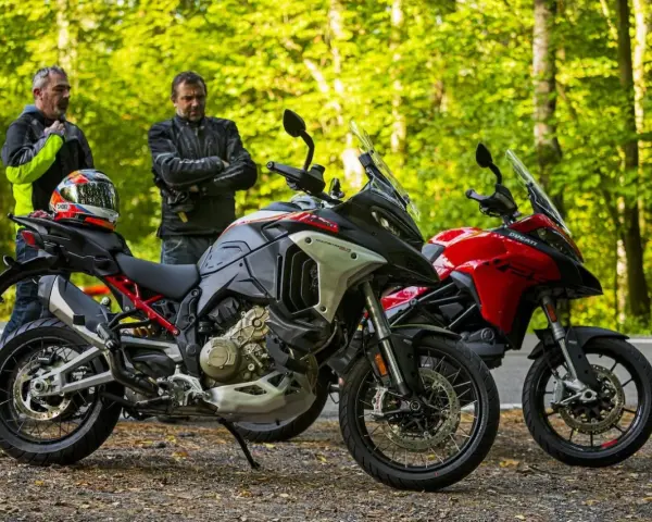 Dwóch motocyklistów z kaskiem na siedzeniu obok dwóch motocykli Ducati Multistrada, szary i czerwony, na tle leśnej drogi.