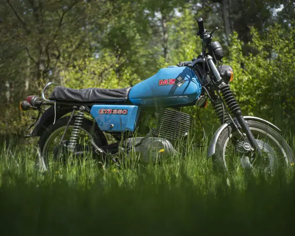 Niebieski motocykl MZ ETZ 250 stojący na trawie, z drzewami w tle.