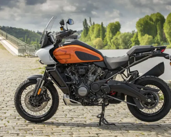 Pomarańczowy Harley-Davidson Pan America gotowy na przygodę, z mostem i zielenią w tle.